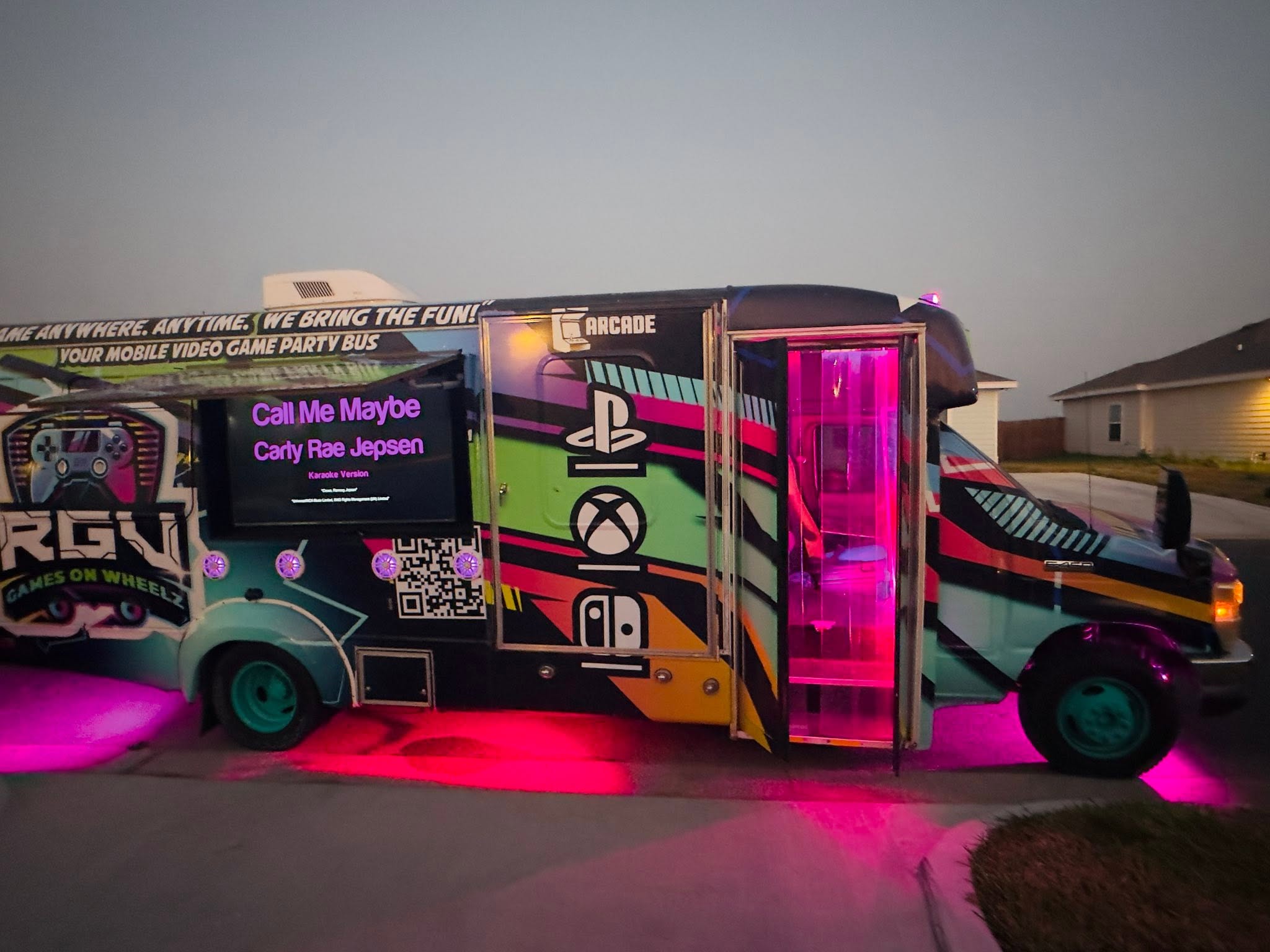The outdoor screen on the side of the RGV Games on Wheelz bus running a karaoke track at dusk, with the magenta LED under-glow lit
