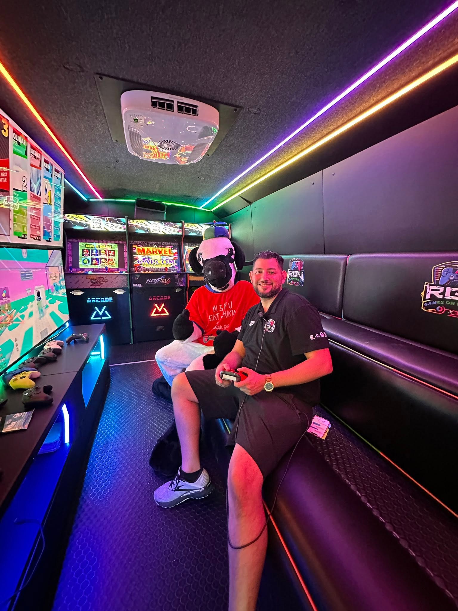 The founder of RGV Games on Wheelz seated with a game controller beside the custom arcade cabinets inside the mobile gaming bus