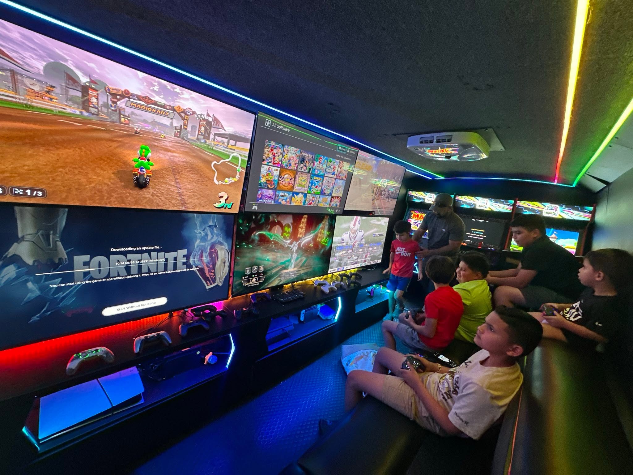 Players at the console stations inside the RGV Games on Wheelz mobile gaming bus with the screen wall lit up